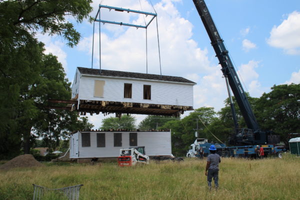 A crane lifts the second story off the a white clapboard two story house.